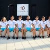 Otto persone sedute con divise bianche e azzurre davanti a un'insegna.