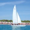 Vela bianca sul mare con spiaggia e ombrelloni sullo sfondo.